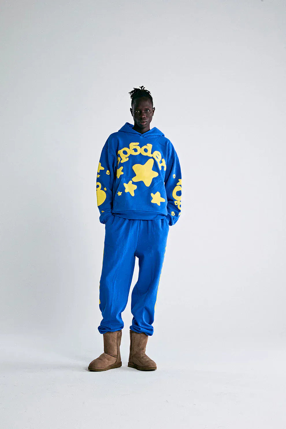 Person stands against a plain background, wearing the Sp5der 440GSM Heavyweight Hoodie in royal blue with yellow star 3D puff print, matching sweatpants, brown boots, and channeling Y2K streetwear vibes.