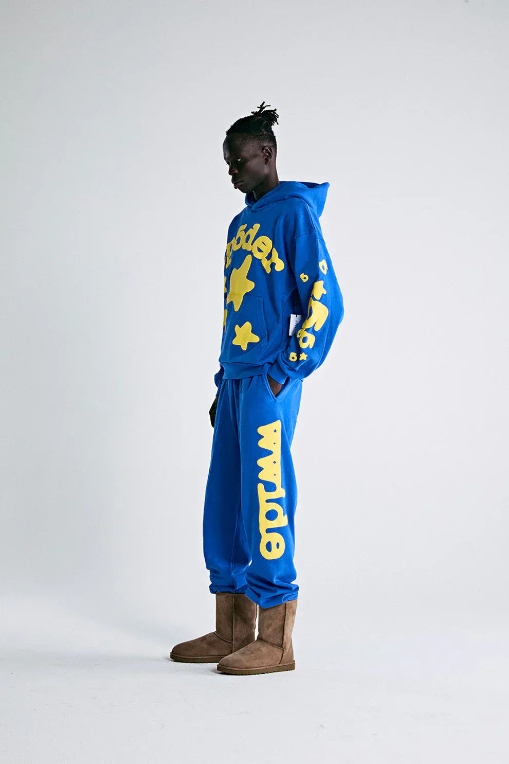 A person stands sideways in the Sp5der 440GSM Heavyweight Hoodie – Royal Blue with Yellow Star 3D Puff Print and matching sweatpants, channeling Y2K streetwear vibes, finished with brown boots.