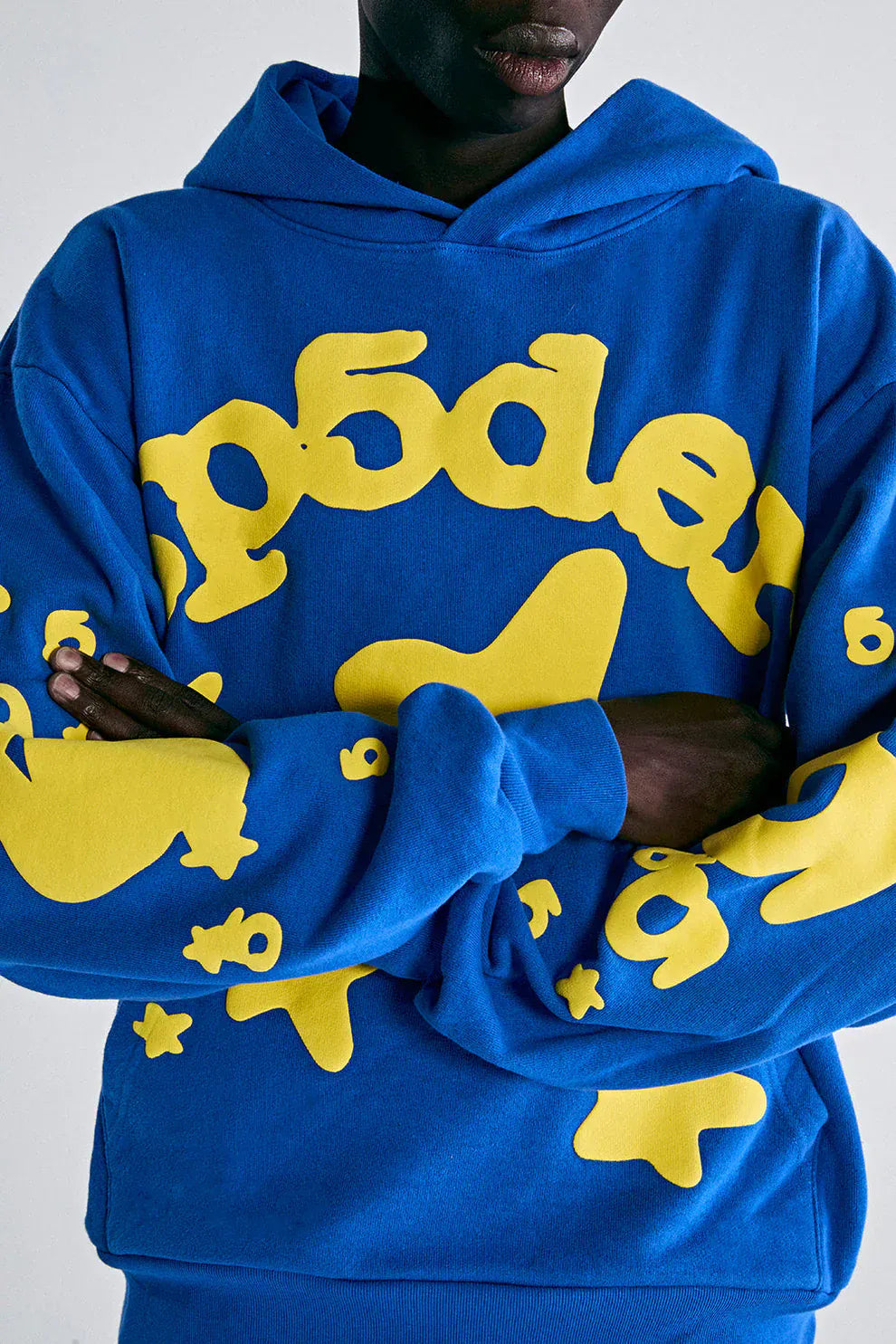 Model wears the Sp5der 440GSM Heavyweight Hoodie in royal blue featuring yellow star 3D puff print, arms crossed against a plain background—ideal for fans of bold Y2K streetwear style.