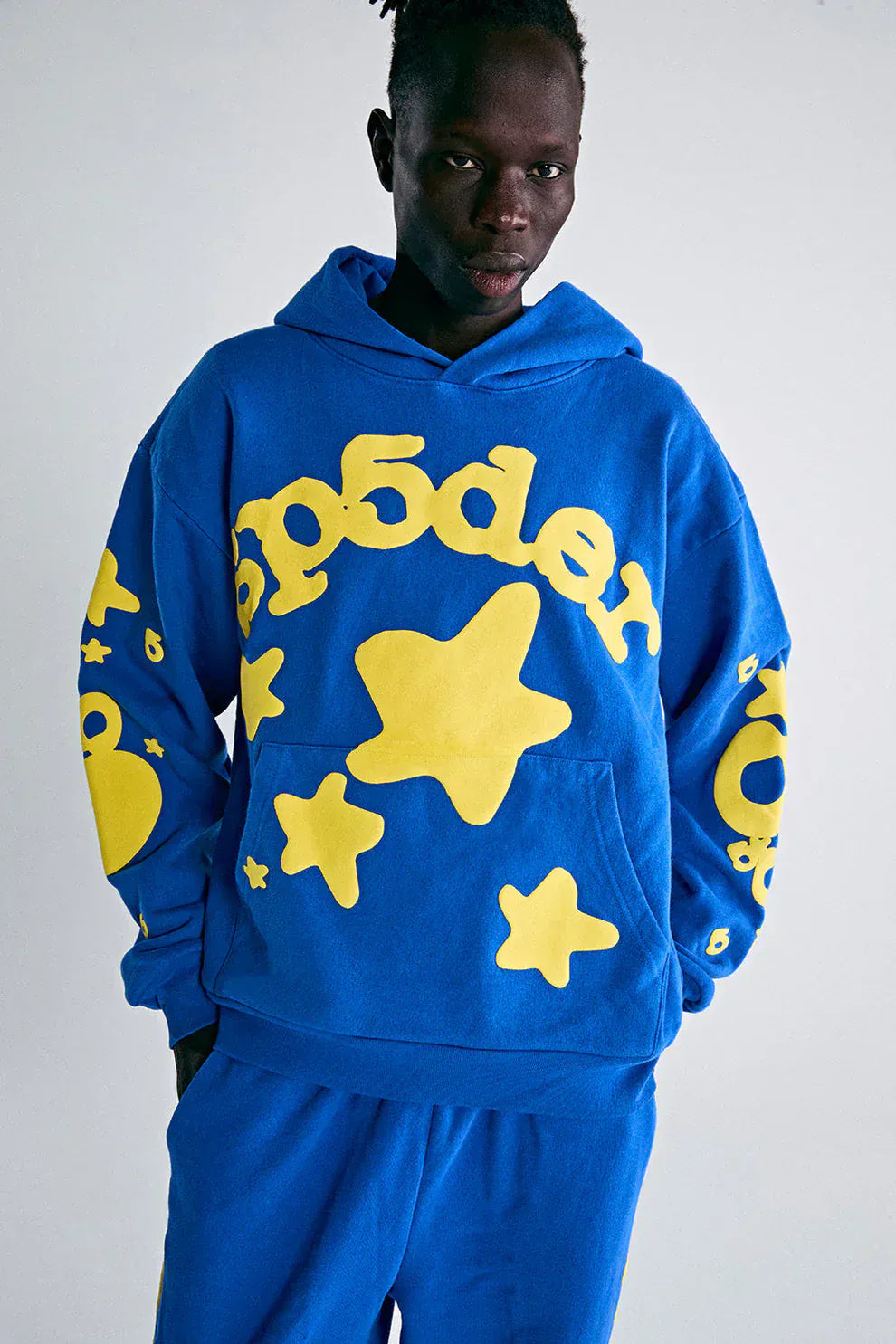 A person stands against a plain light background wearing the Sp5der 440GSM Heavyweight Hoodie in royal blue, featuring large yellow star 3D puff prints and Y2K streetwear accents, styled with matching pants.