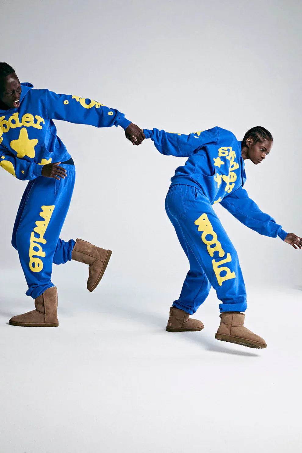 Two people in matching Sp5der 440GSM Heavyweight Hoodies in royal blue with yellow star 3D puff print hold hands; one pulls the other forward. Both wear light brown boots, standing against a plain white background.