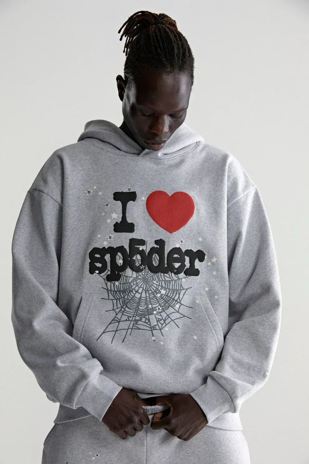 A person in the Sp5der “I Love Sp5der” Hoodie – Grey 440GSM Heavyweight Oversized Streetwear Pullover, featuring an "I ♥ spider" and web graphic, stands against a plain light background and looks down.