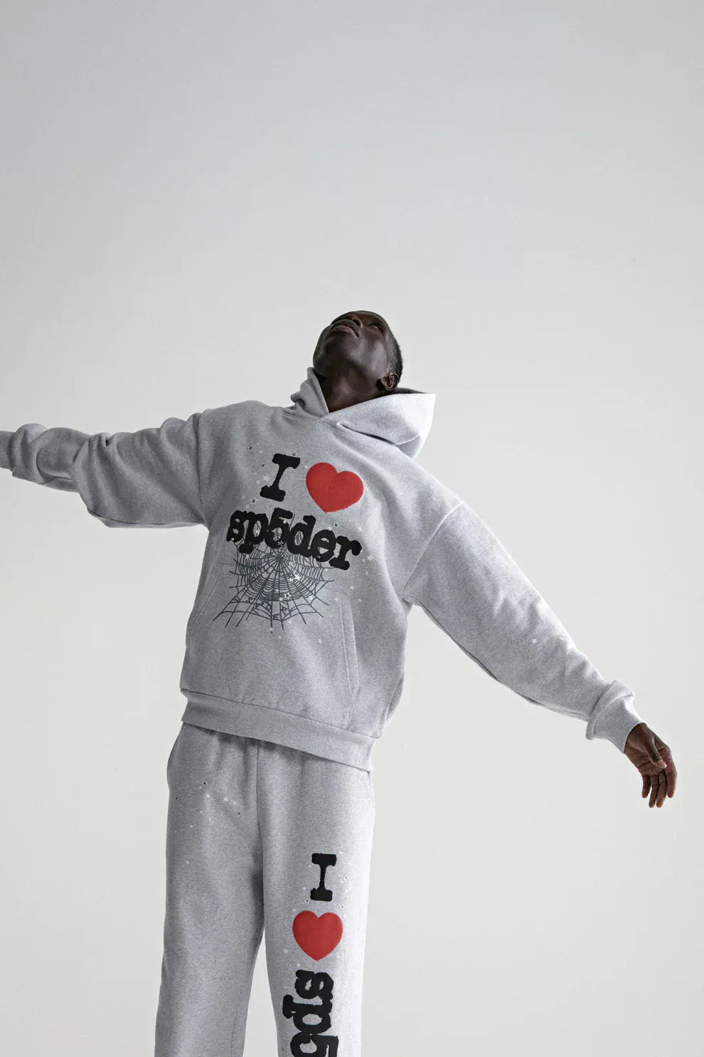 A person wears the Sp5der “I Love Sp5der” Hoodie – Grey 440GSM heavyweight oversized streetwear pullover with “I love Sp5der” text and spider web graphic, standing arms outstretched against a plain light background.