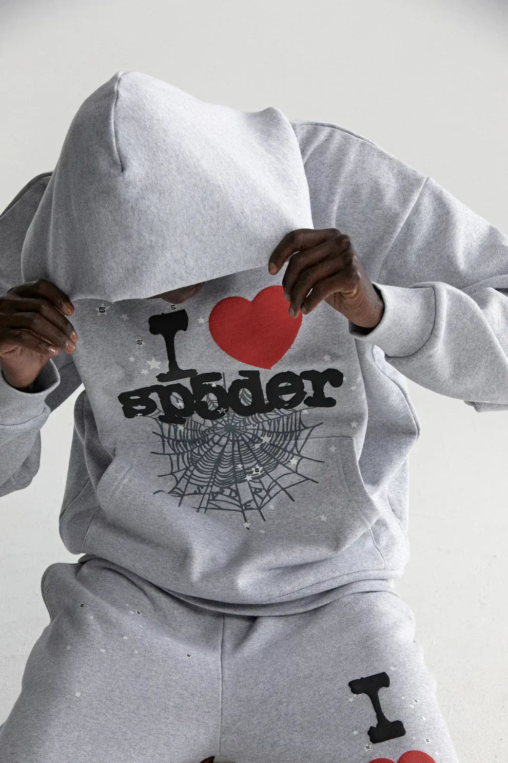 A person wearing the Sp5der “I Love Sp5der” Hoodie – Grey 440GSM Heavyweight Oversized Streetwear Pullover with an "I ♥ spider" and web graphic pulls the hood over their face while seated against a plain background.