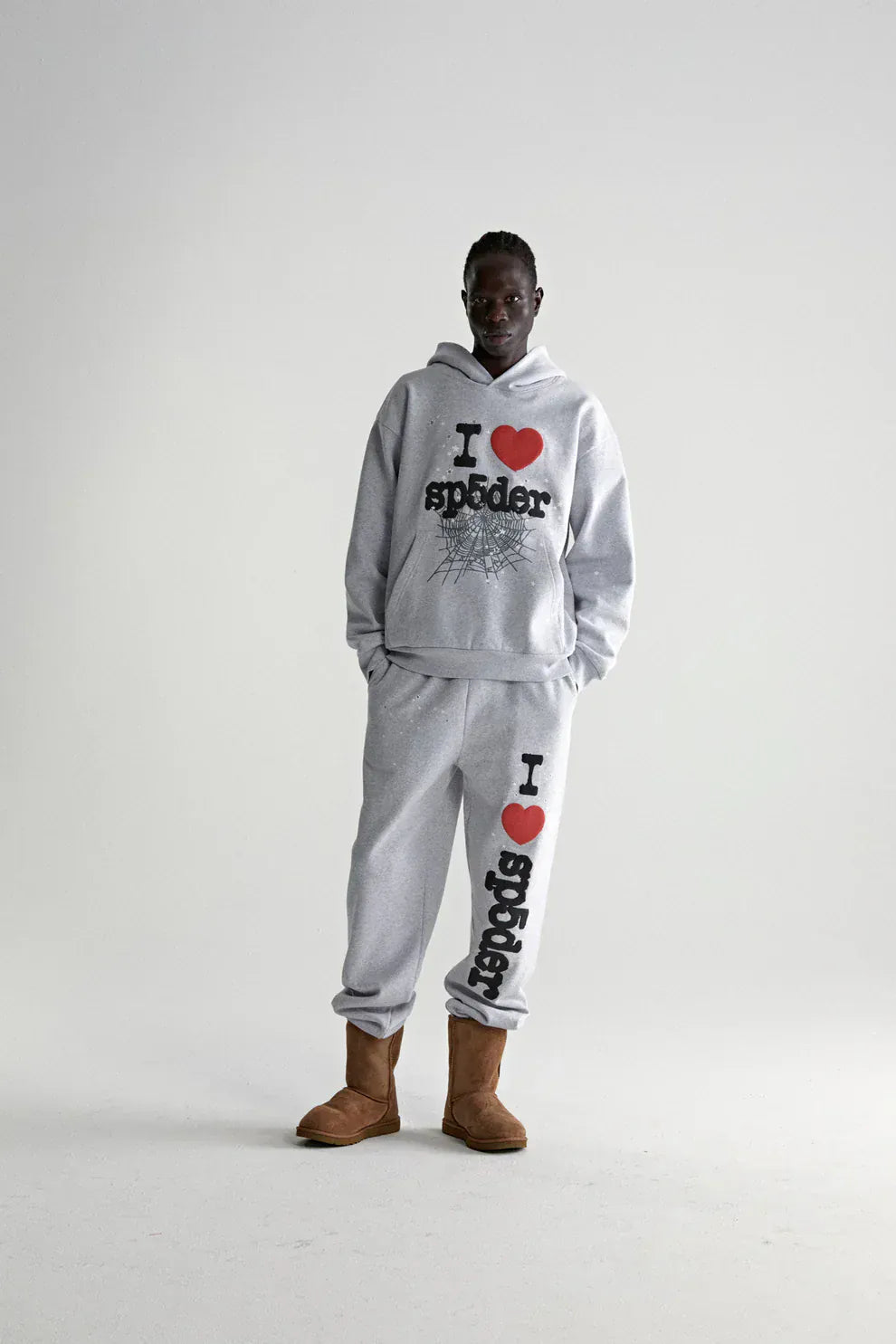 A person stands against a plain background wearing a Sp5der “I Love Sp5der” Hoodie – Grey 440GSM Heavyweight Oversized Streetwear Pullover, paired with sweatpants and brown boots, hands in pockets for a relaxed Y2K streetwear vibe.