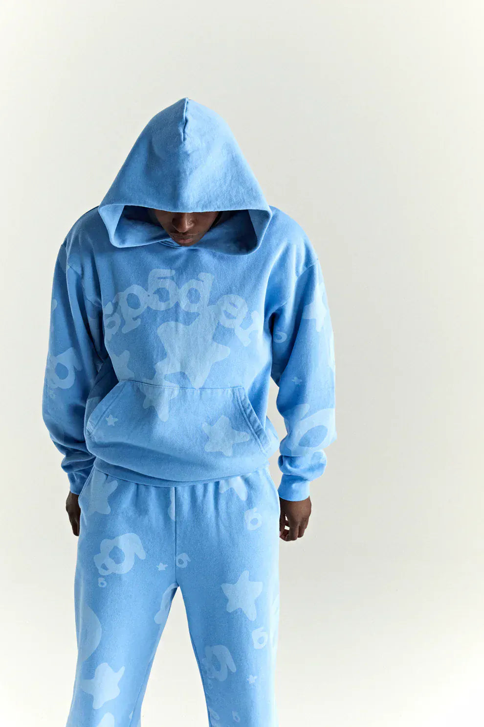 A person in a Sp5der Star Hoodie – Baby Blue 440GSM Heavyweight Oversized Streetwear Pullover with matching sweatpants, featuring star details and Y2K vibes, stands against a plain background, hood up and looking down.