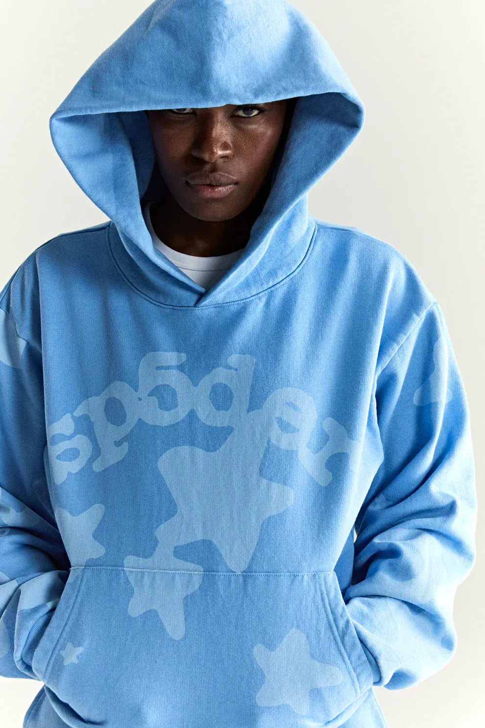 A person wearing the Sp5der Star Hoodie – Baby Blue 440GSM Heavyweight Oversized Streetwear Pullover, featuring white printed stars and text on the front, stands against a plain backdrop for a cool Y2K vibe.