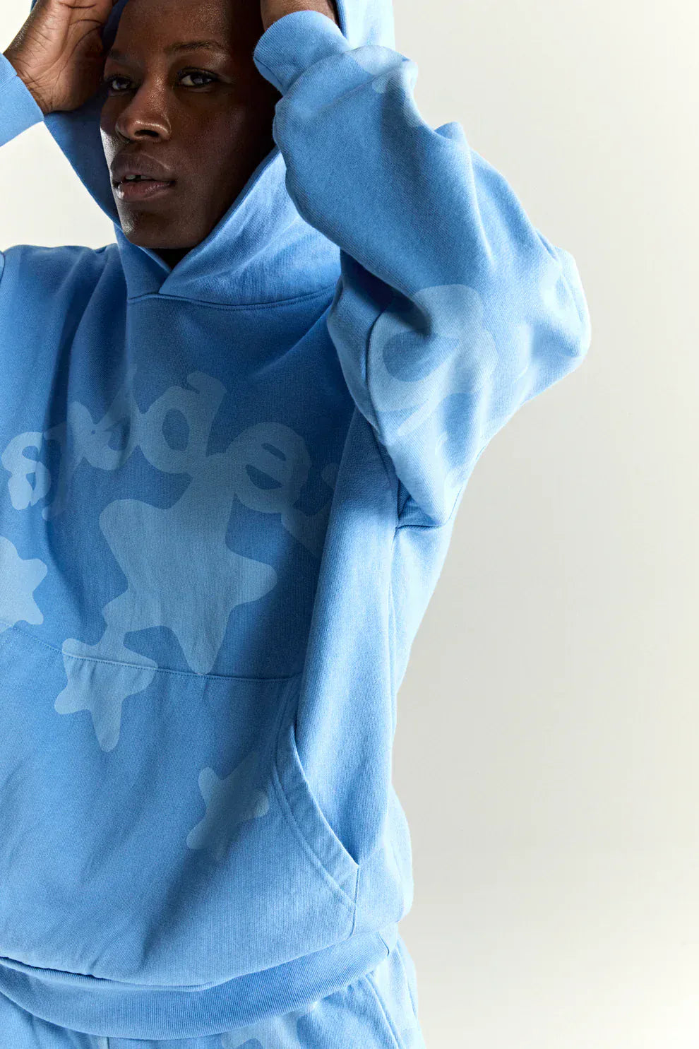 A person in a Sp5der Star Hoodie – Baby Blue 440GSM Heavyweight Oversized Streetwear Pullover adjusts the hood, wearing matching sweatpants and showcasing the hoodie’s large abstract pattern.