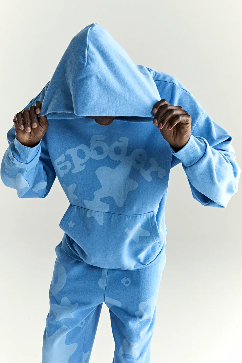 Wearing the Sp5der Star Hoodie – Baby Blue 440GSM Heavyweight Oversized Streetwear Pullover, a person lifts the hood to partly cover their face, channeling bold Y2K streetwear vibes against a plain background.