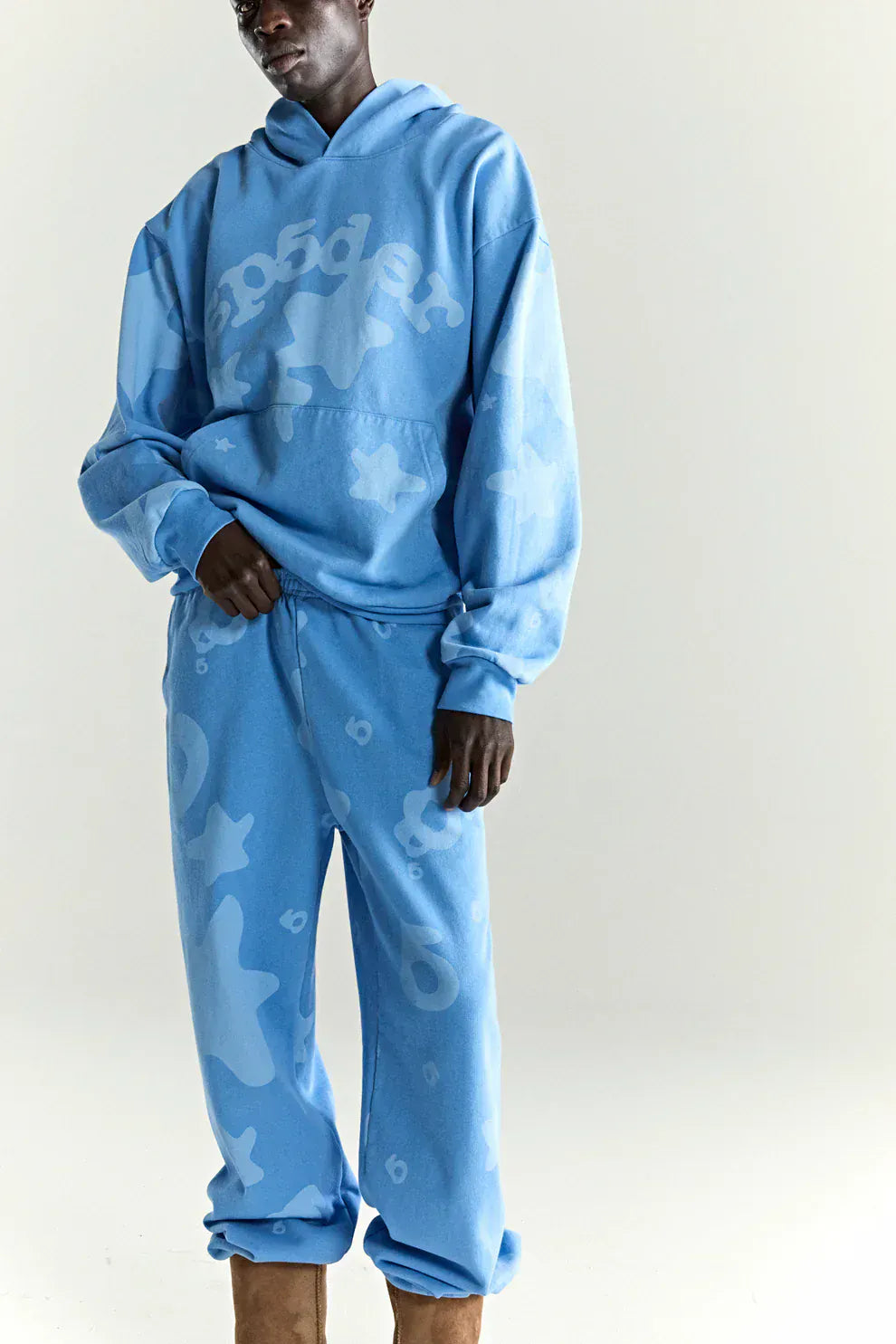 A person wears a Sp5der Star Hoodie – Baby Blue 440GSM Heavyweight Oversized Streetwear Pullover with matching sweatpants featuring abstract patterns and text, embodying a bold Y2K vibe against a plain light background.