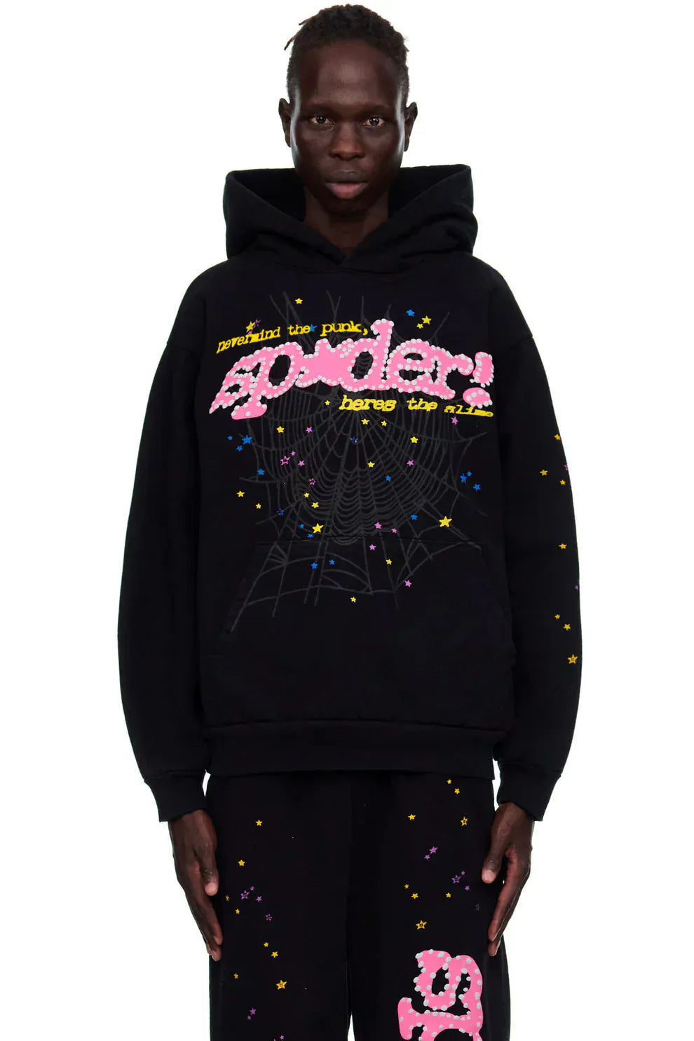 A person wears the Sp5der “Hares the Slime” Hoodie—black 440GSM heavyweight spider-web streetwear pullover with pink and yellow “spider!” print—and matching black fleece pants, standing against a white background.