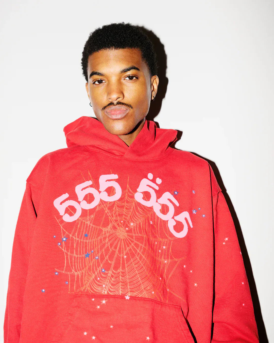 A person stands against a white background wearing the Sp5der Worldwide Red Angel Number 555 Hoodie—a red and pink oversized streetwear pullover with spiderweb details, triple 555 print on the front, and crafted from 440GSM heavyweight cotton fleece.