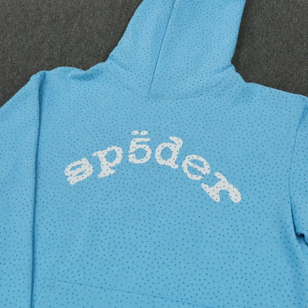 The Sp5der Baby Blue Rhinestone All-Over Arch Logo Hoodie features small black dots and a white, stylized "sp5der" print across the back. This trendy streetwear item is shown on a dark fabric surface.