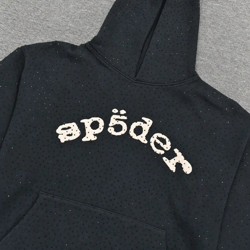 The Sp5der Black Rhinestone Hoodie Cream Arch Logo Cotton features subtle rhinestone embellishments and a stylized “sp5der” arch logo on the chest, laid flat on a gray surface to showcase its streetwear style.