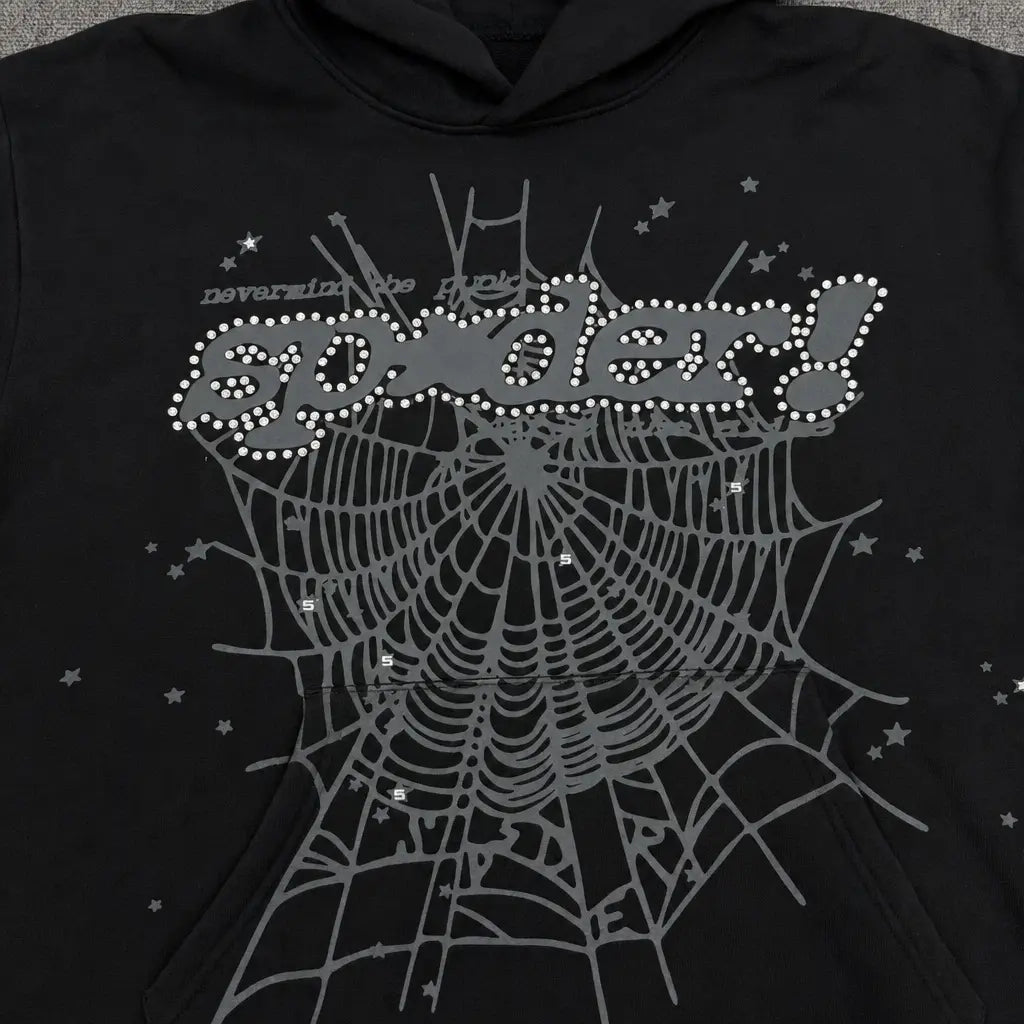 The Sp5der Black Rhinestone Spiderweb Graphic Hoodie features a large gray rhinestone spiderweb, white star accents, a bold dotted "spider!" logo on the chest, and “Devarjuna the Holy” text above for standout streetwear style.