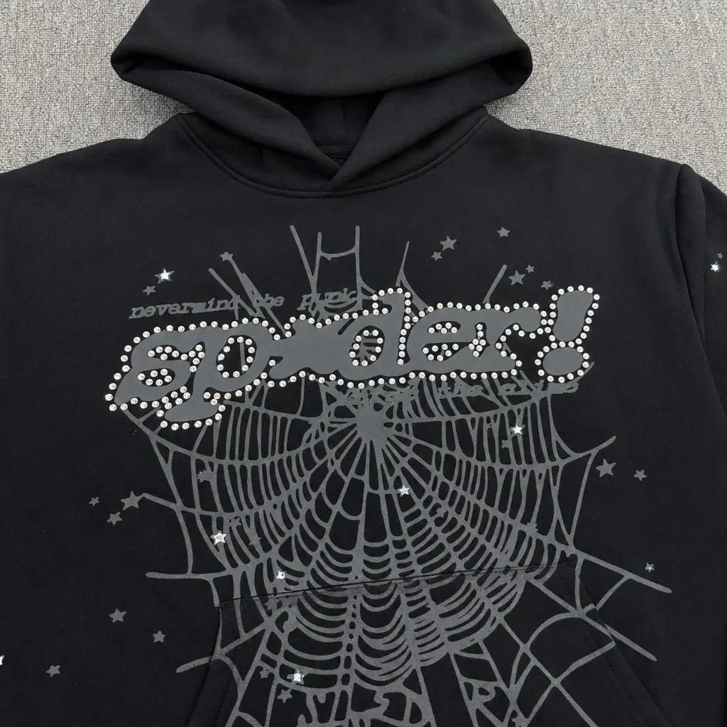 The Sp5der Black Rhinestone Spiderweb Graphic Hoodie showcases a silver spiderweb with sparkling accents, rhinestone logo, bold dotted "spider!" text on the chest, and floating stars and "never in the hell" above―ideal for streetwear style.