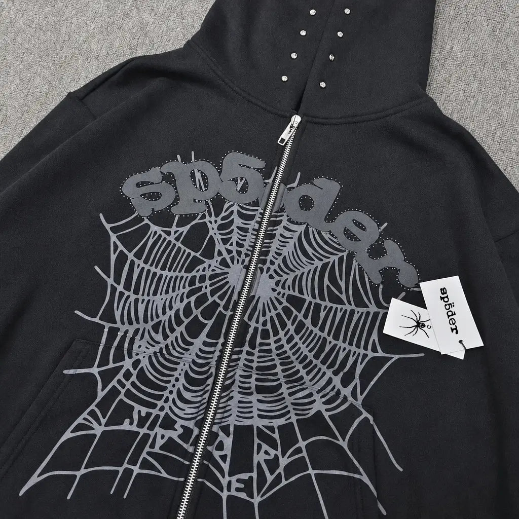 The Sp5der Black Zip-Up Hoodie with Spiderweb Graphic features silver-studded hood details, a bold gray web design on the front, "Sp5der" printed above, and a matching spider tag on the zipper for streetwear appeal.