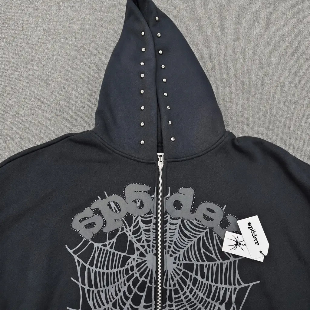 The Sp5der Black Zip-Up Hoodie with Spiderweb Graphic features silver studs on the hood, a spiderweb graphic with "sp5der" on the chest, and a white hoodie tag with a spider illustration hanging from the zipper.