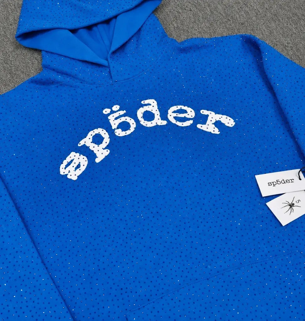 The Sp5der Blue Rhinestone All-Over Arch Logo Hoodie is a blue streetwear hoodie with silver rhinestones and “sp?der” in white, stylized text across the chest, plus a tag featuring the same text and a spider illustration.