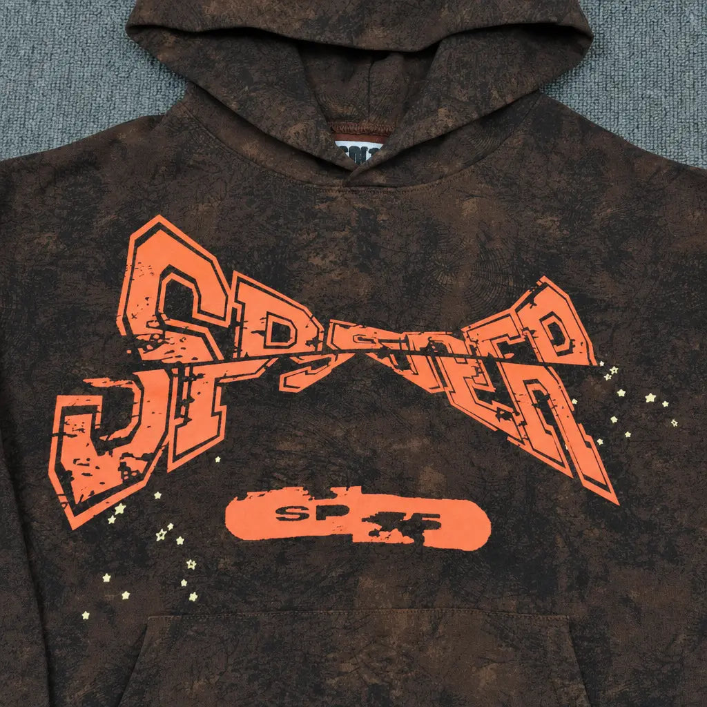The Sp5der Brown Tie-Dye Hoodie with Orange Logo Print in streetwear style features bold orange SPSTER text, star accents, and a skateboard graphic, displayed on a textured gray surface.