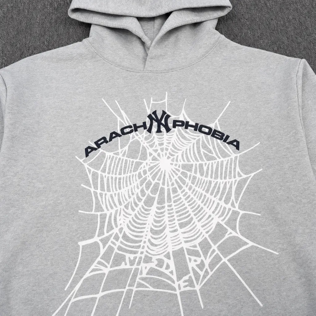 The Sp5der Arachnophobia NY Spiderweb Graphic Hoodie Grey features a striking white spiderweb and curved black ARACHNOPHOBIA text on the front, laid flat on a dark textured surface for a modern streetwear appeal.