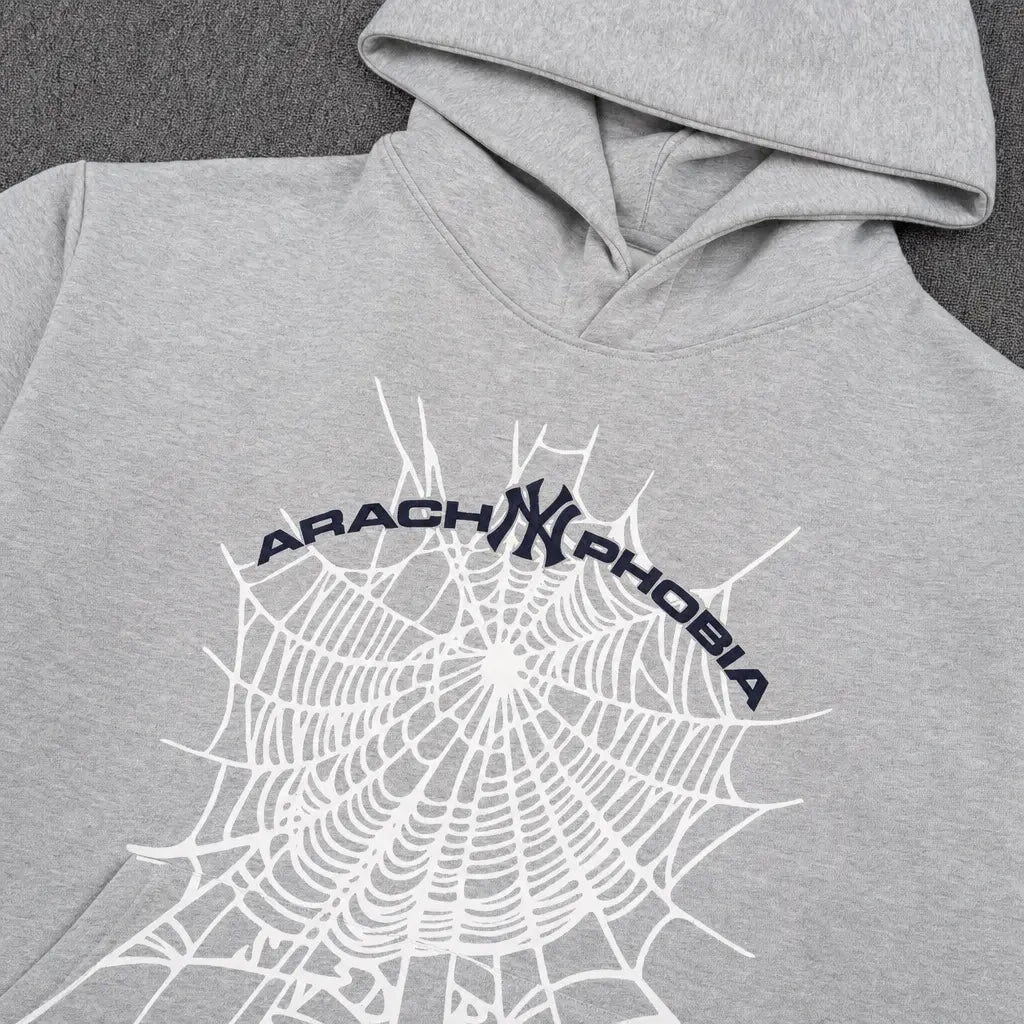 The Sp5der Arachnophobia NY Spiderweb Graphic Hoodie Grey features a bold spiderweb design, dark blue ARACHNOPHOBIA text above, and a stylized NY logo at the center―a standout piece for any streetwear collection.