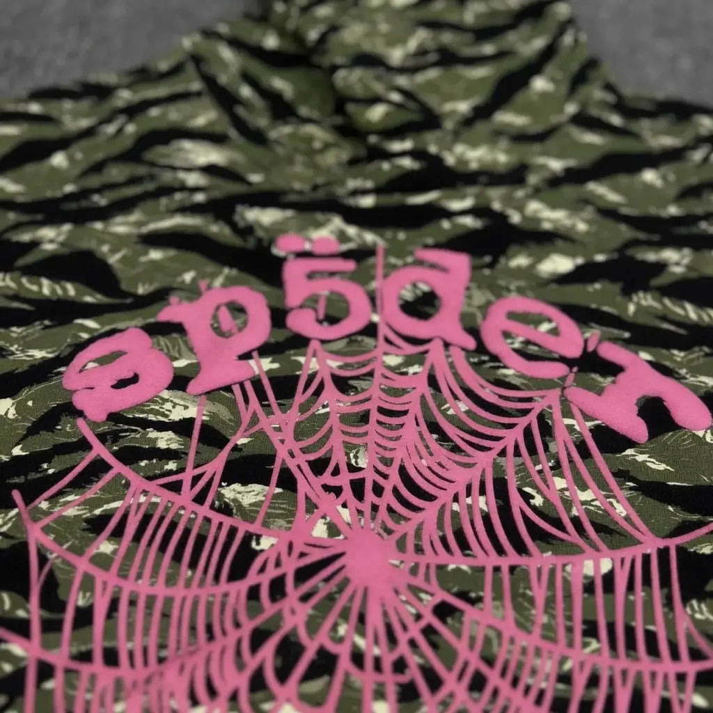 The Sp5der Tiger Camo Hoodie features green tiger camo fabric with "spider" in pink stylized text above a pink spiderweb logo, making it a bold and eye-catching streetwear statement.
