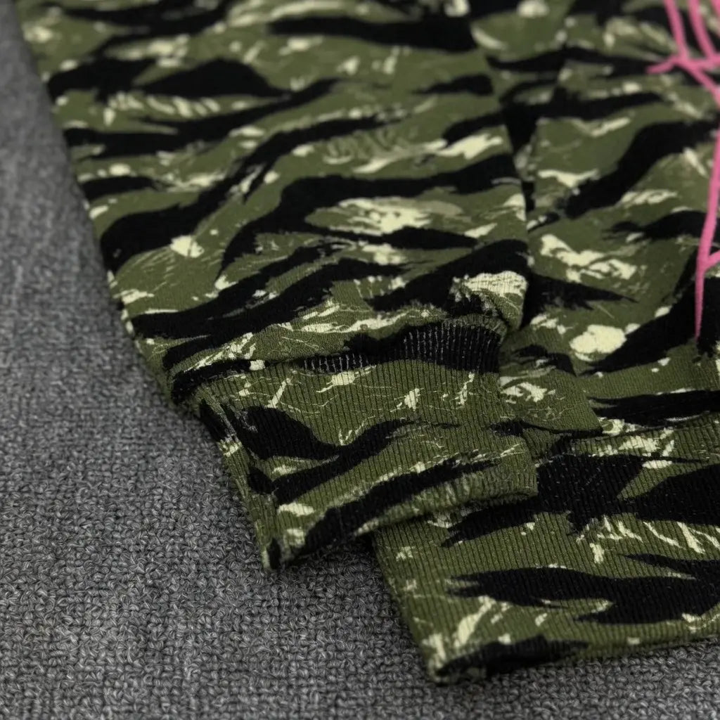 Close-up of the Sp5der Tiger Camo Hoodie with Pink Spiderweb Logo in green camo, featuring ribbed cuffs and a few visible pink threads, displayed on a textured grey surface.