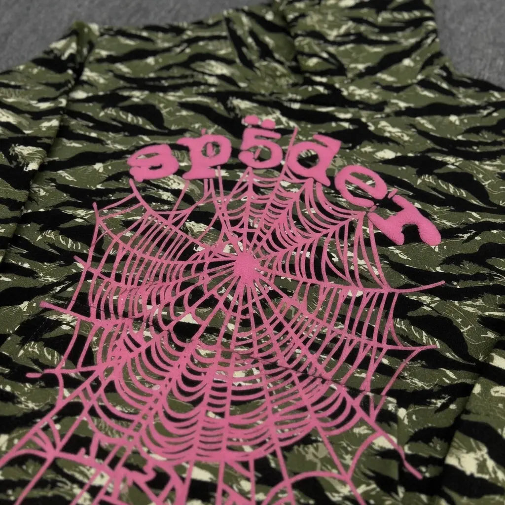 The Sp5der Tiger Camo Hoodie with Pink Spiderweb Logo features green tiger camo, black camouflage, a bold pink spiderweb graphic, and “spēder” in reverse above it―ideal for standout streetwear style.