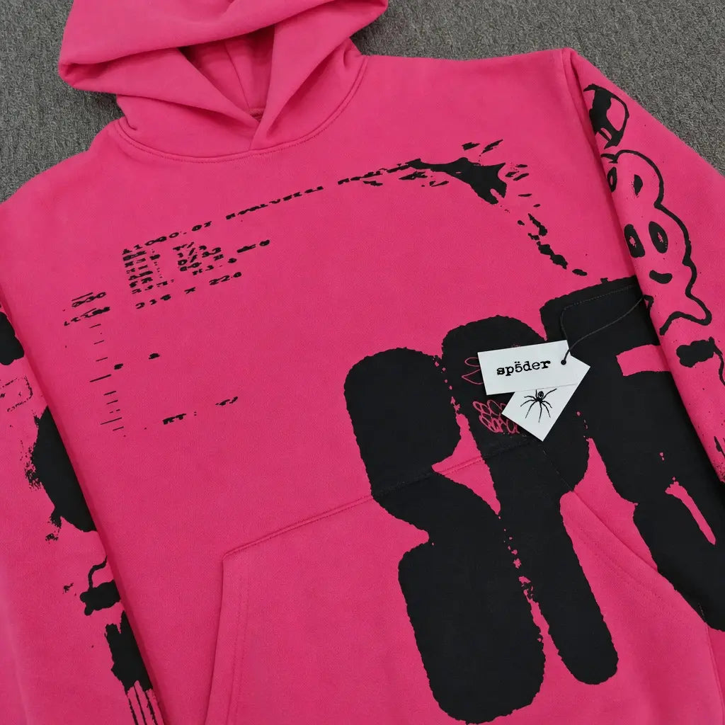 The Sp5der Hot Pink Hoodie Black Graffiti Text Print features bold graphics and a signature "sp5der" tag on the front pocket, laid flat on a grey surface―an ideal choice for any streetwear collection.