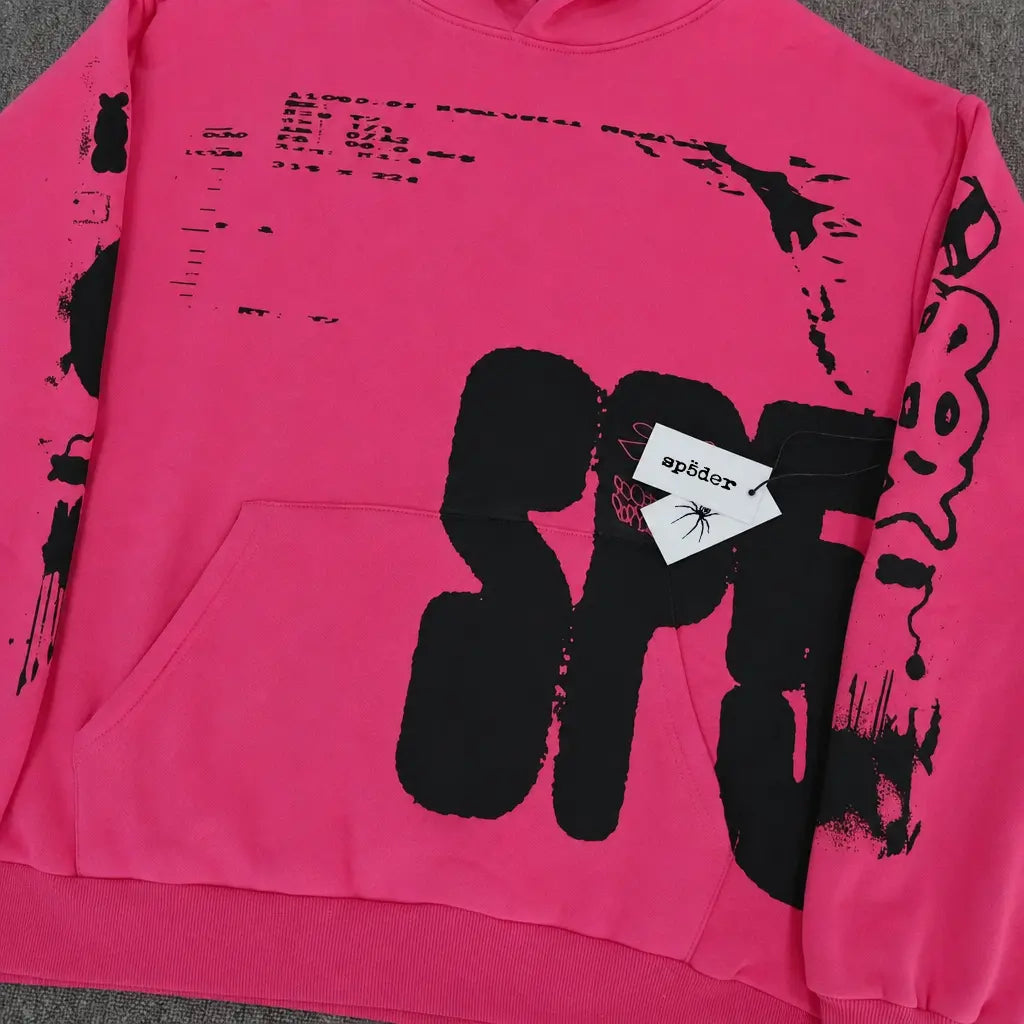 The Sp5der Hot Pink Hoodie Black Graffiti Text Print features abstract black lettering, a white sp5der tag with a spider graphic on the front pocket, and is displayed flat on a gray surface.
