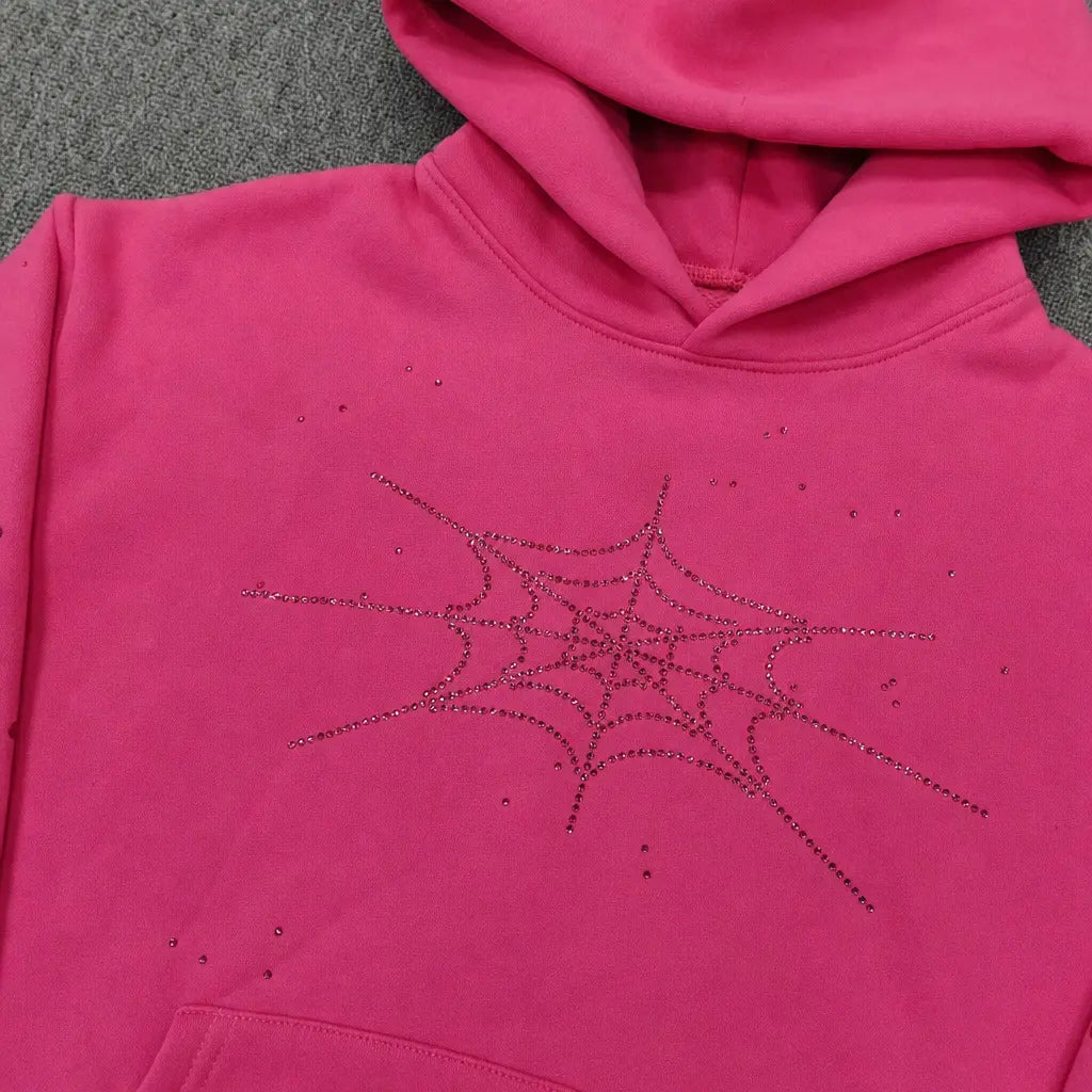 The Sp5der Pink Rhinestone Spiderweb Crystal Hoodie features a sparkling spiderweb design on hot pink, displayed on a gray textured surface―an eye-catching addition to any streetwear pullover collection.