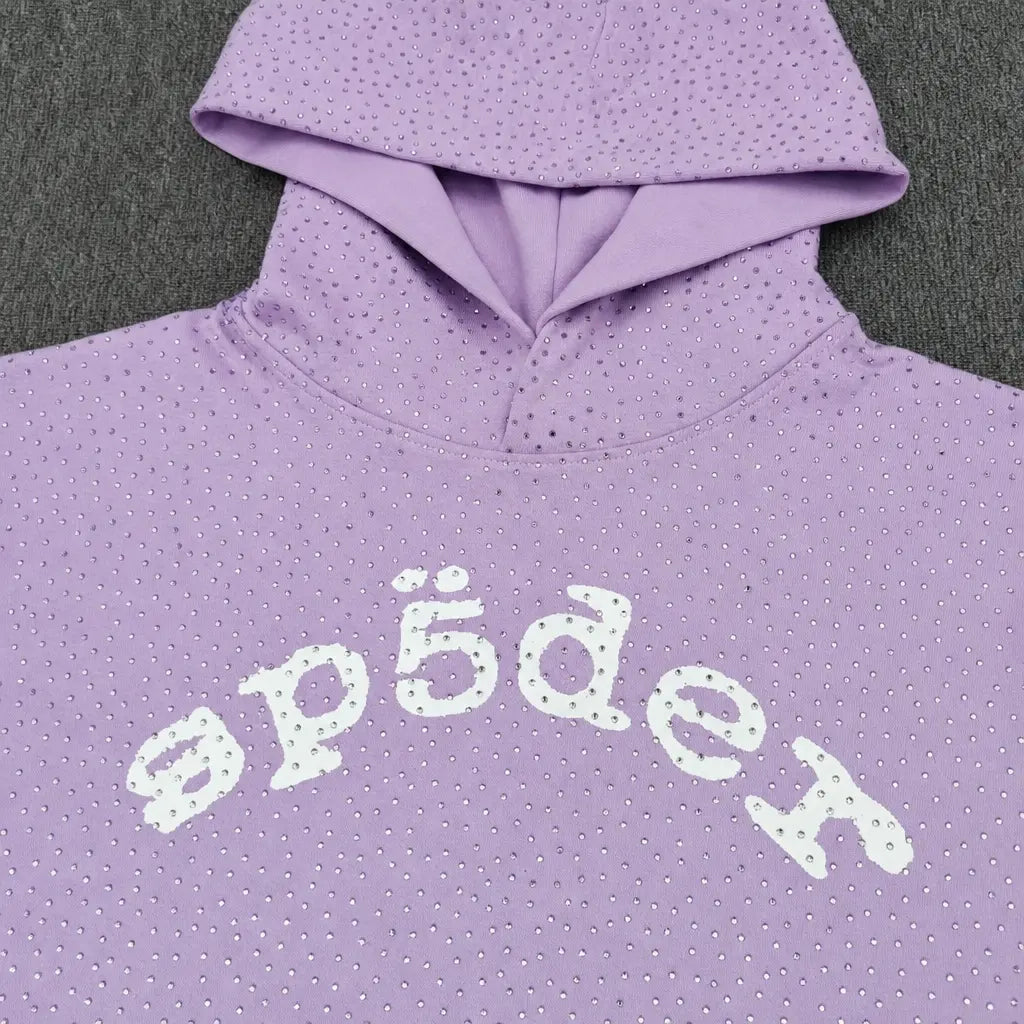 The Sp5der Rhinestone Arch Logo Light Purple Hoodie features small rhinestones and a white "sp5der" arch logo across the chest, shown on a dark gray surface―a standout streetwear essential.