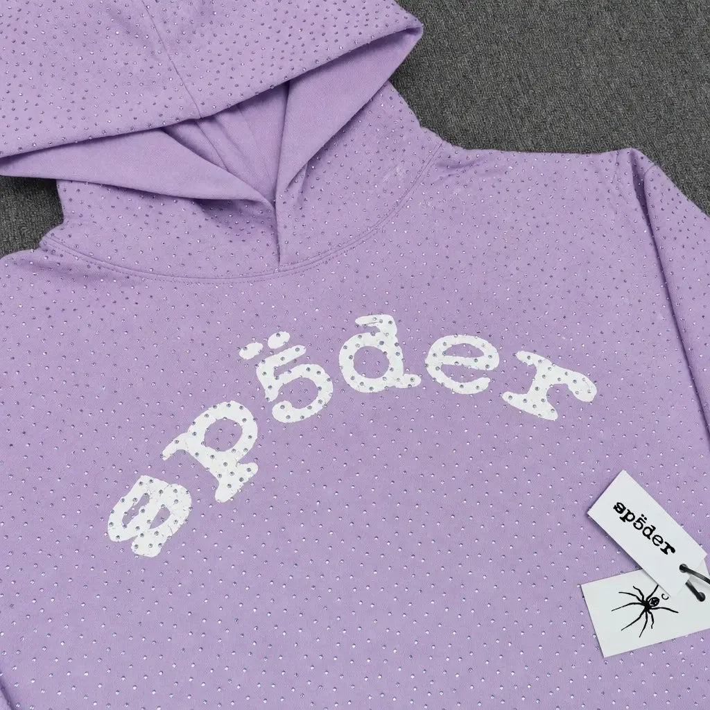 The Sp5der Rhinestone Arch Logo Light Purple Hoodie features sparkling rhinestones and a white textured "sp5der" logo on the chest, plus a branded tag with a spider graphic, all showcased on a dark gray surface.