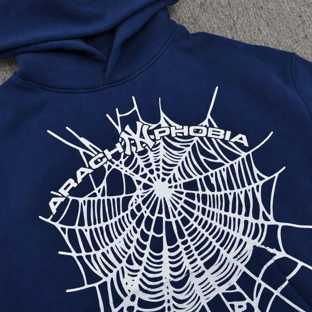 The Sp5der Arachnophobia Navy Blue Spiderweb Print Hoodie features a bold white spiderweb and "ARACHNOPHOBIA" across the back, shown laid flat on a textured gray surface.