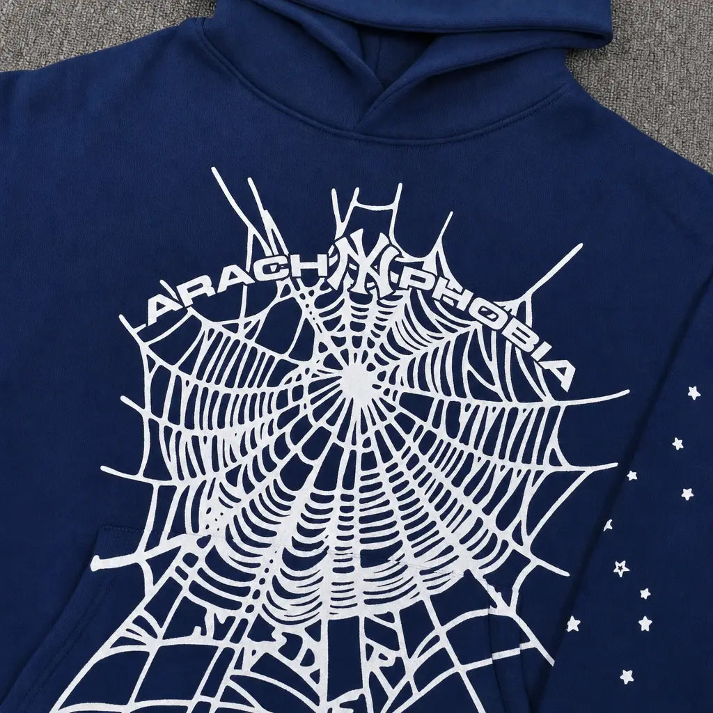 The Sp5der Arachnophobia Navy Blue Spiderweb Print Hoodie features a large white spiderweb graphic with "ARACHNOPHOBIA" curved above and small white stars on one sleeve.