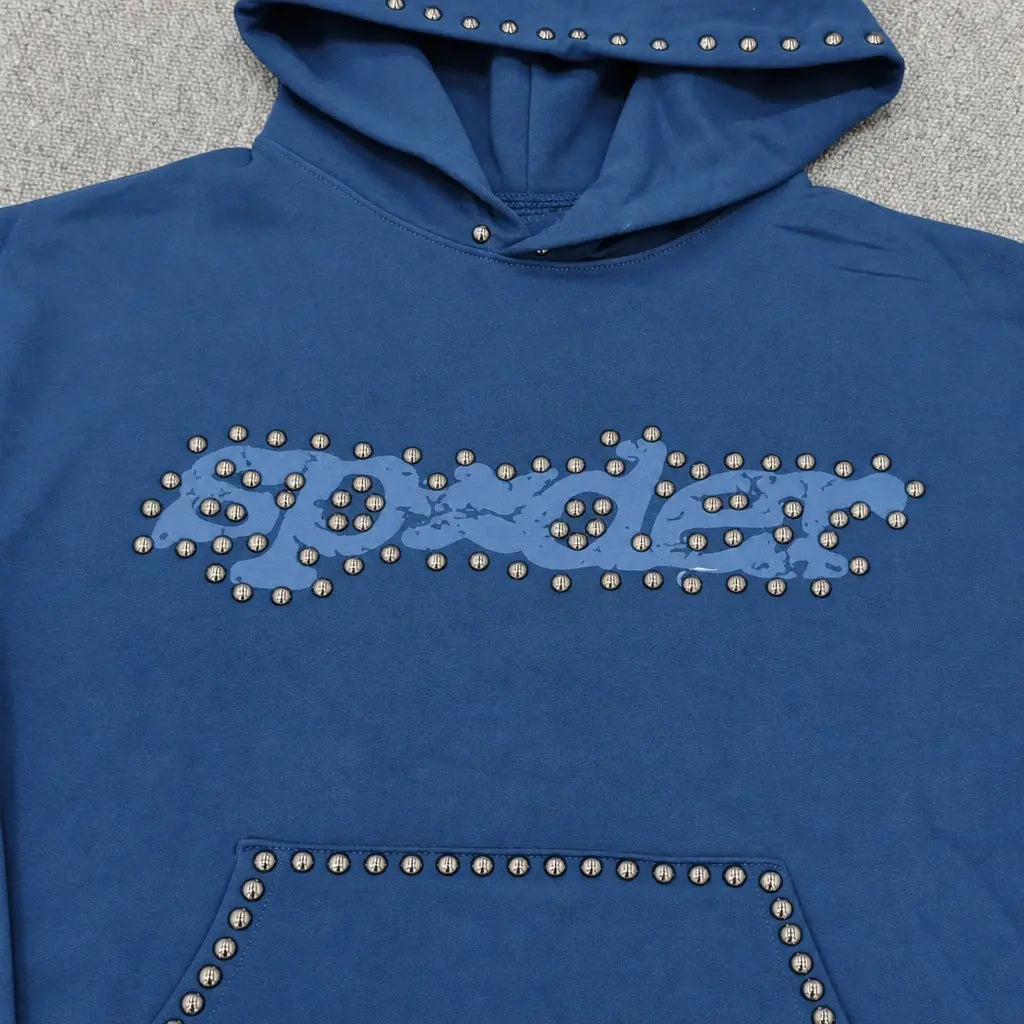 The Sp5der Navy Blue Studded Logo Hoodie 100% Cotton features a studded chest logo outlined with silver studs, plus matching silver studs on the hood and front pocket. The fabric is displayed on a gray surface.
