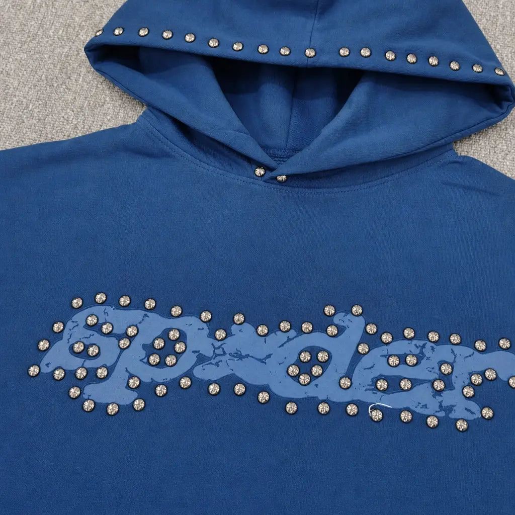 The Sp5der Navy Blue Studded Logo Hoodie 100% Cotton features silver studs outlining the logo and hood, with a distressed "spider" graphic, displayed on a light gray surface.