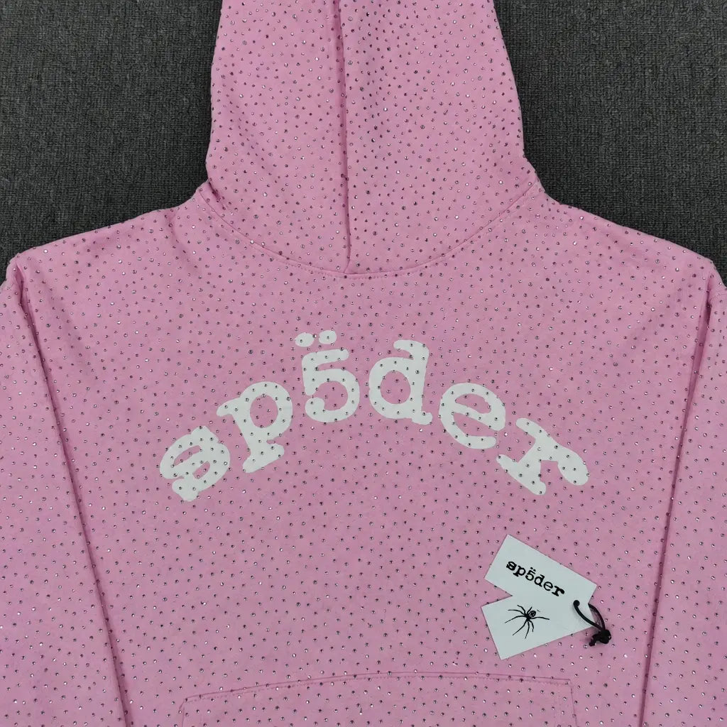 The Sp5der Pink Rhinestone All-Over Arch Logo Hoodie features small black dots, bold white “sp5der” text across the chest, and a tag with the sp5der logo and a spider graphic, making it a stylish streetwear piece.
