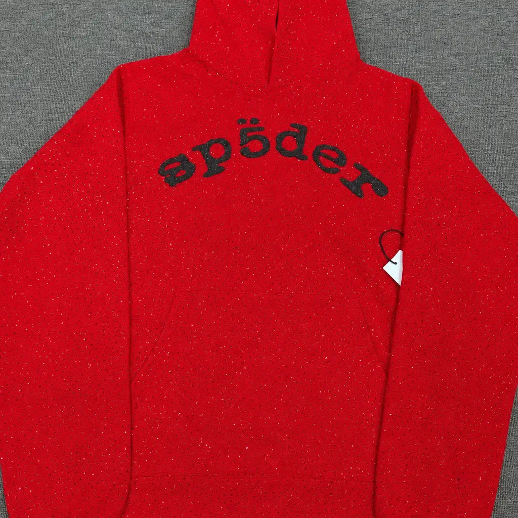 The Sp5der Red Glitter Knit Hoodie with Black Arch Logo features a hood, black stylized "sp5der" lettering across the chest, and a white side tag. This streetwear hoodie is displayed flat on a gray surface.