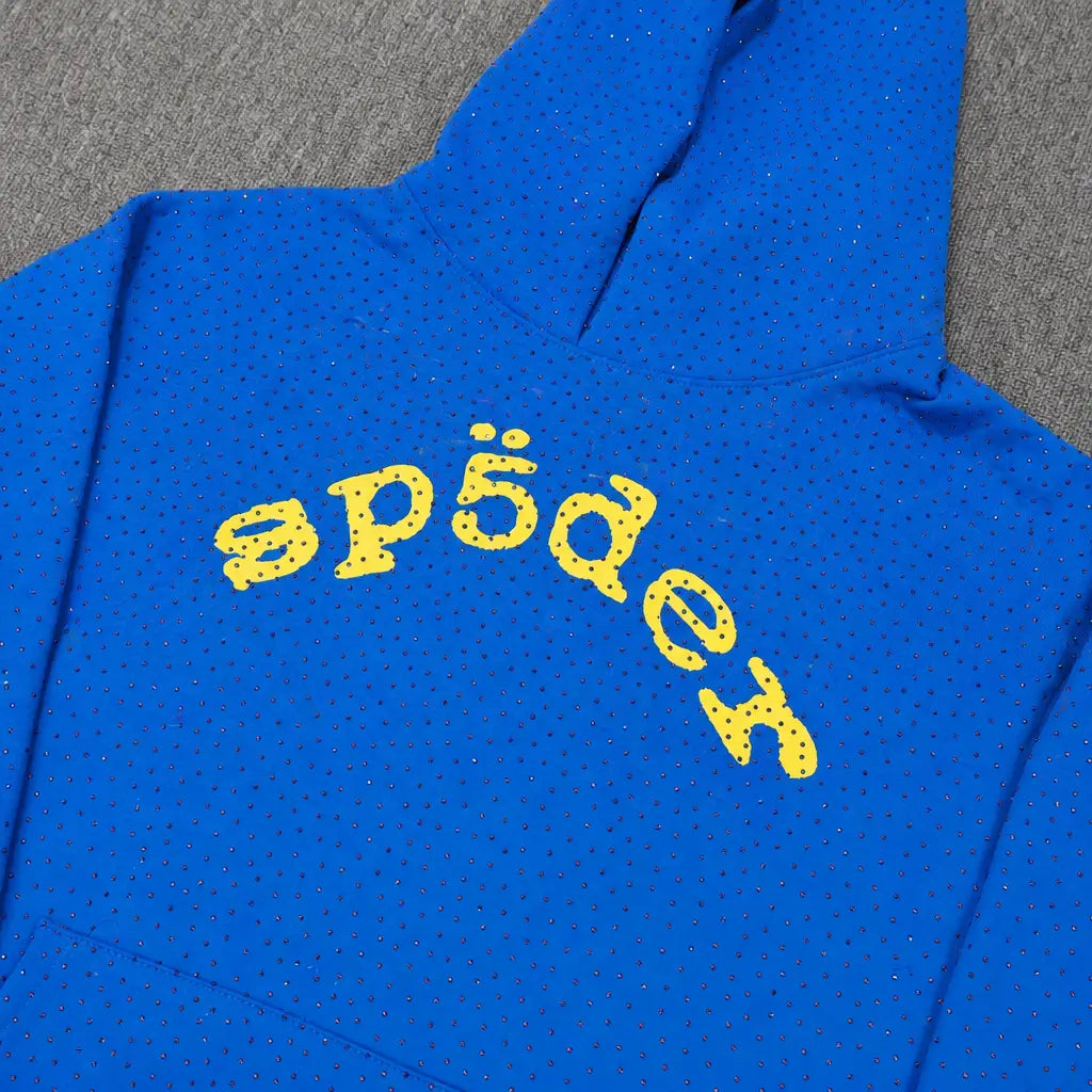 The Sp5der Royal Blue Rhinestone Hoodie features a yellow arch logo across the chest and small black dots, laid flat on a gray surface.