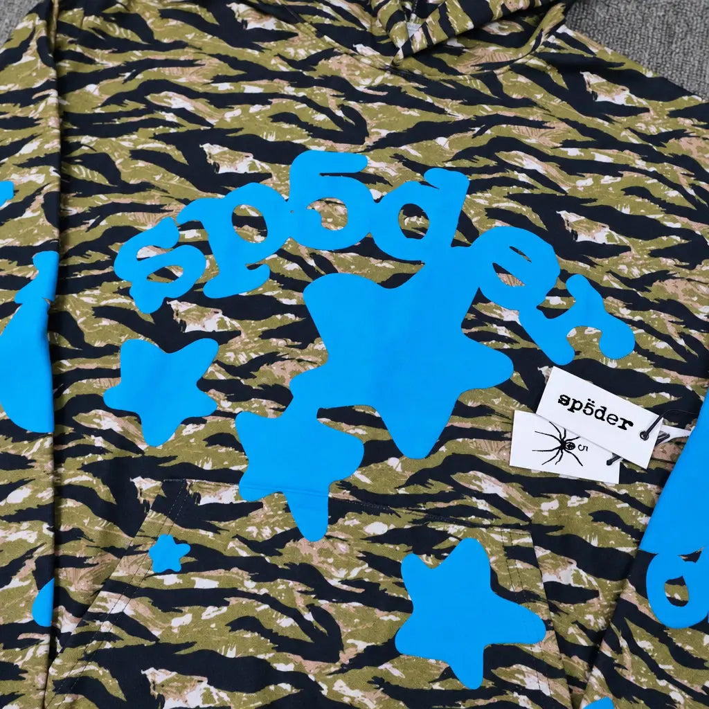 The Sp5der Tiger Camo Blue Star Print Logo Hoodie features a green and black tiger camo pattern, blue stars, bold blue “sp5der” text, and two white tags―one with the sp5der logo and a spider graphic.