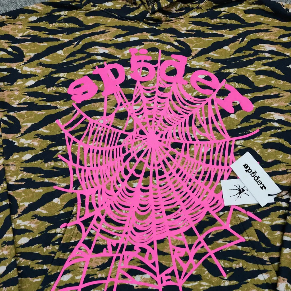 The Sp5der Tiger Camo Hoodie features a streetwear-inspired camouflage pattern, a large pink spiderweb print with "sp5der" in pink text, and two attached white tags―one displaying the sp5der logo and a black spider graphic.