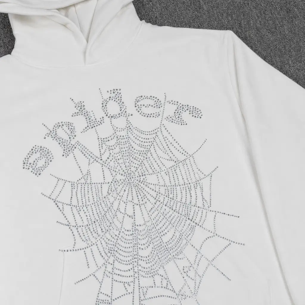 The Sp5der White Rhinestone Spiderweb Crystal Logo Hoodie features a crystal spiderweb and "spider" logo in rhinestones, laid flat on a gray surface―a bold choice for standout streetwear.