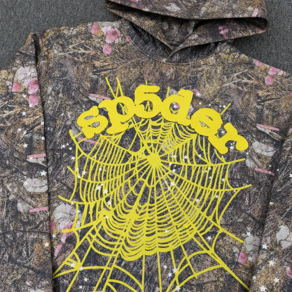 The Sp5der Woodland Camo Floral Web Graphic Hoodie features a camo pattern, bold yellow “spōder” lettering, and a striking spiderweb graphic―delivering standout streetwear style to your collection.