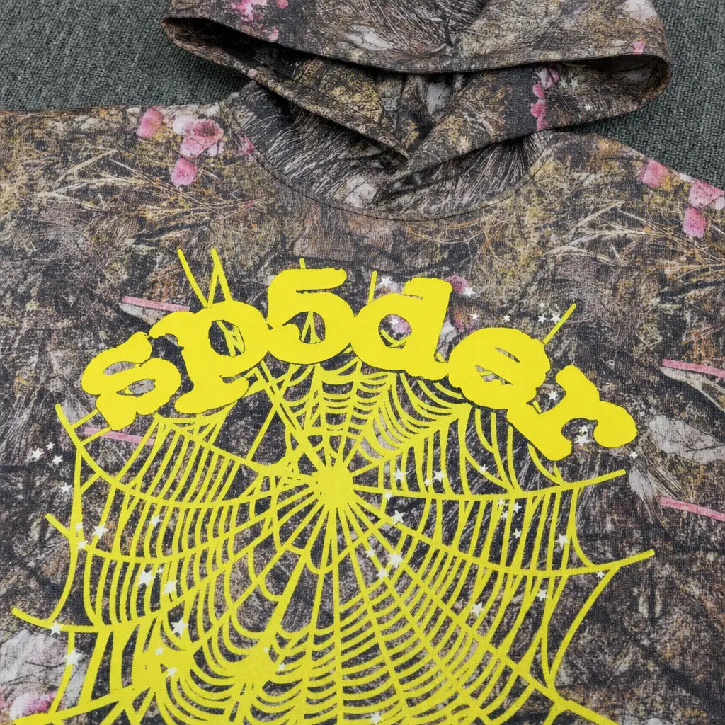 The Sp5der Woodland Camo Floral Web Graphic Hoodie features a striking yellow web graphic and "sp5der" lettering on the front, merging standout urban style with a unique woodland camo and floral pattern.