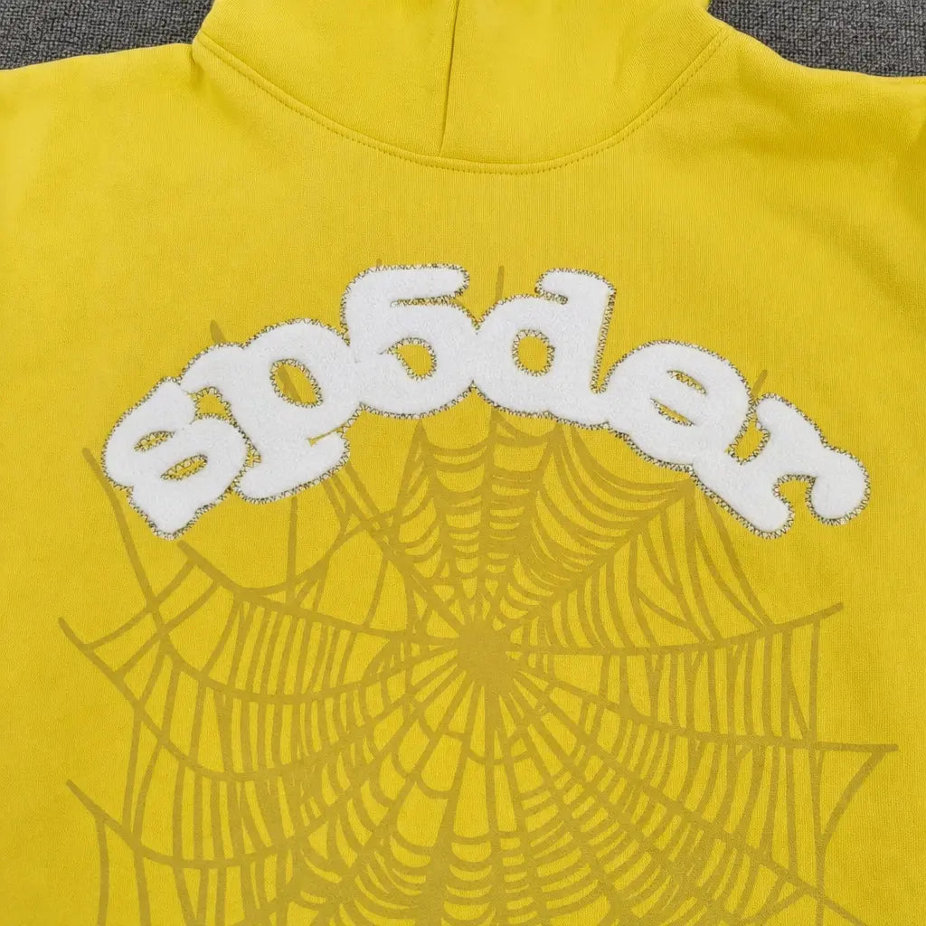 The Sp5der Yellow OG Web Graphic Pullover Hoodie features yellow fabric with "sp5der" in white fuzzy letters above a bold yellow spiderweb graphic, making it a standout streetwear staple.