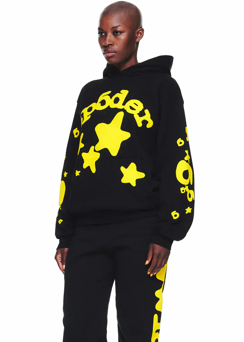 Wearing the Black and Yellow Spider Hoodie with Star Lettering Print, a comfort fit pullover with bold yellow star and circular designs plus “Poder” text, paired with black pants for everyday streetwear style.