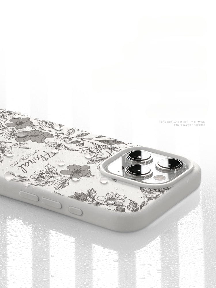 An iPhone 16 Pro Max case, adorned with a white floral pattern, rests on a reflective, wet surface. The camera lenses of the phone can be seen, accentuating the case's design. Text on the case reads "Dirty tolerant without yellowing." Experience full 360° protection and style with this Floral Silicone Protective Cover featuring a shockproof triple-layer design and anti-fingerprint coating.