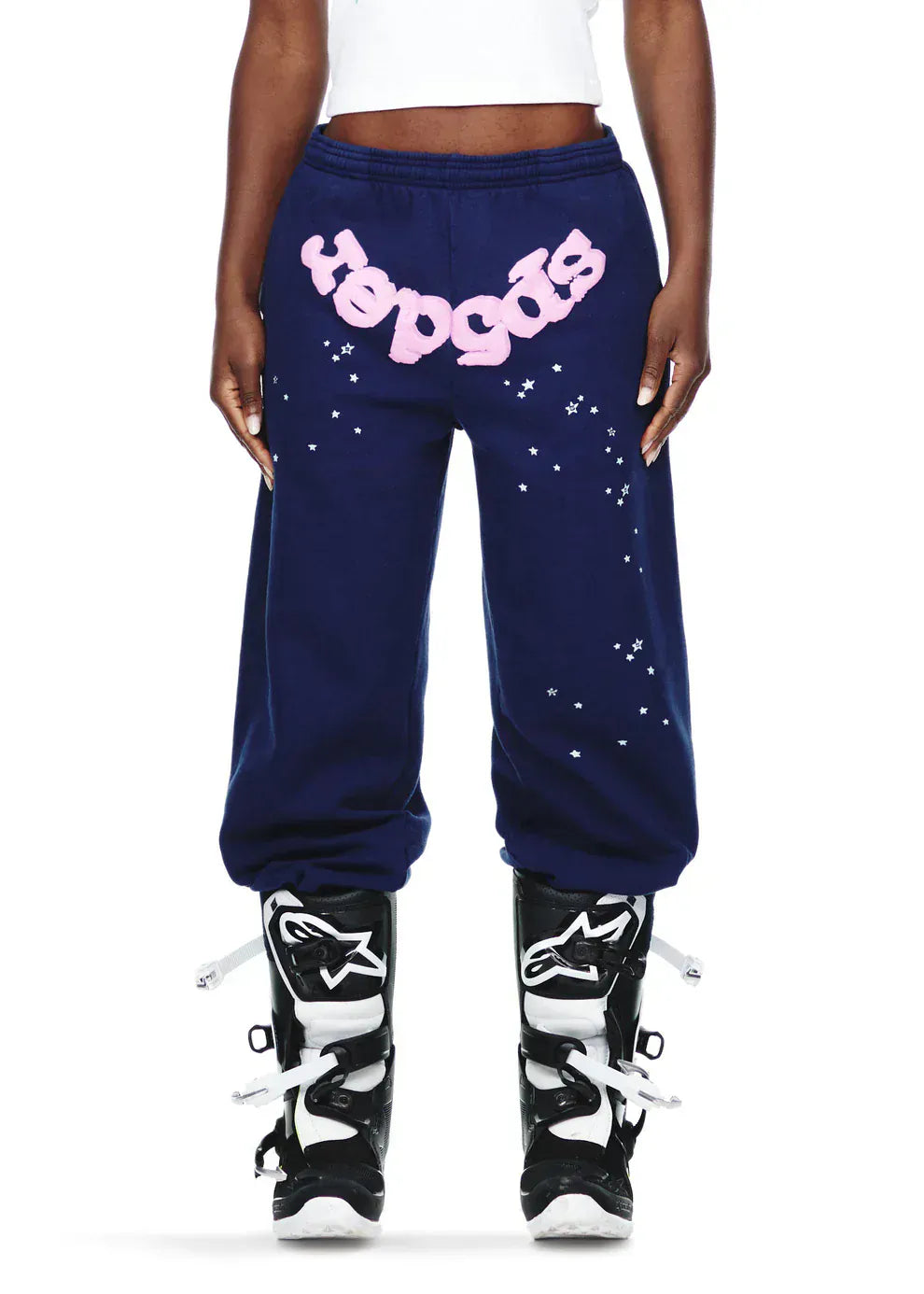 Someone rocks Sp5der Sweatpants—unisex navy blue cotton joggers with a pink front logo and star print—with motocross boots and a white crop top for bold streetwear energy.